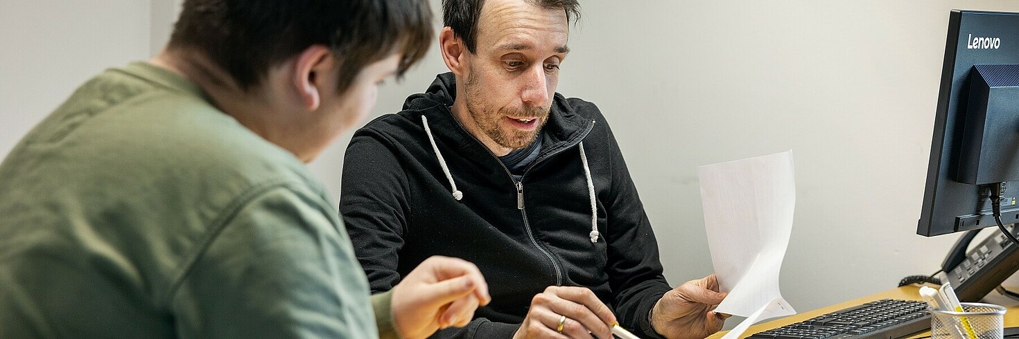 Ein Mann und ein Jugendlicher sitzen an einem Schreibtisch. Der Mann erklärt etwas.